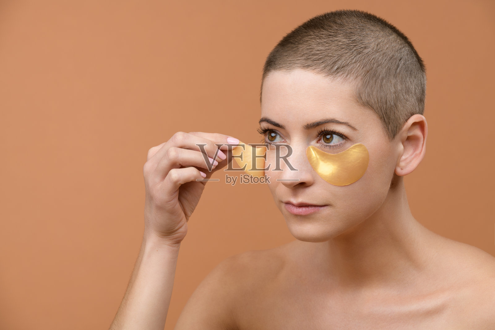 迷人的年轻女子使用黄金胶原眼膜。中年白人妇女的照片剃光头和健康的皮肤隔离在米色背景。美容产品的概念。照片摄影图片