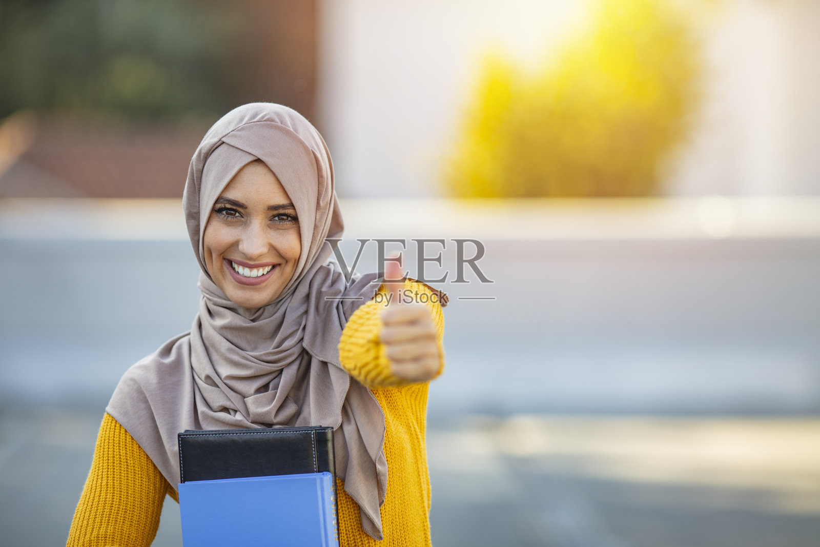 快乐微笑的穆斯林女学生与书显示拇指向上的手势。照片摄影图片