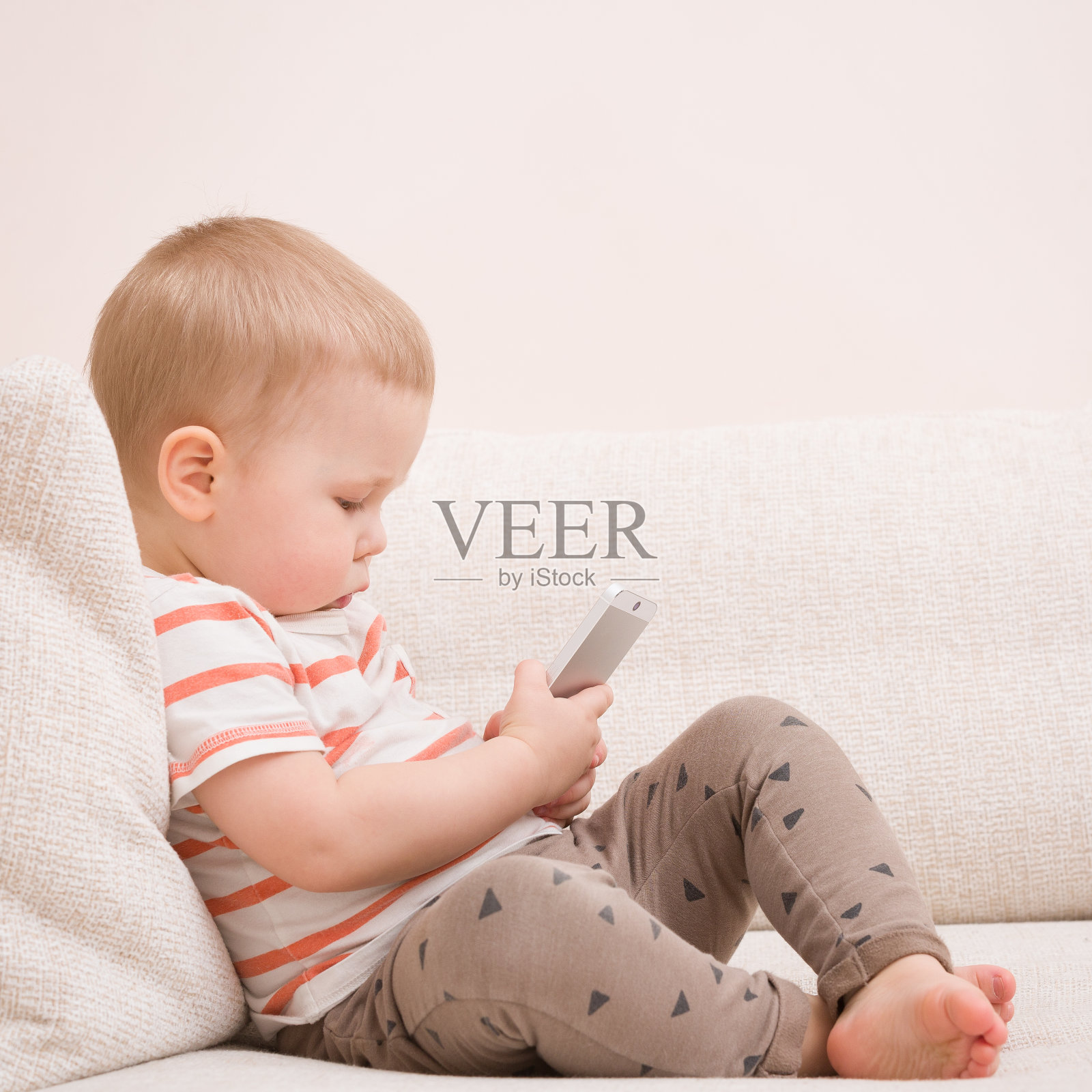 可爱的小男孩坐在客厅的沙发上玩手机。孩子正在学习如何使用智能手机。男孩在电话上发短信。-科技和生活理念。照片摄影图片