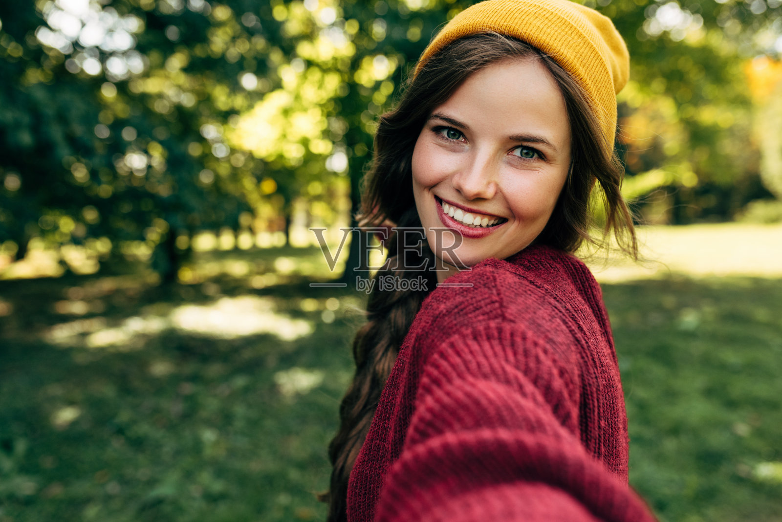 美丽的年轻金发女人笑容灿烂，健康的白牙齿，穿着一件红色的毛衣，和一顶黄色的帽子，在自然背景上自拍。快乐的女性在智能手机上自拍。照片摄影图片