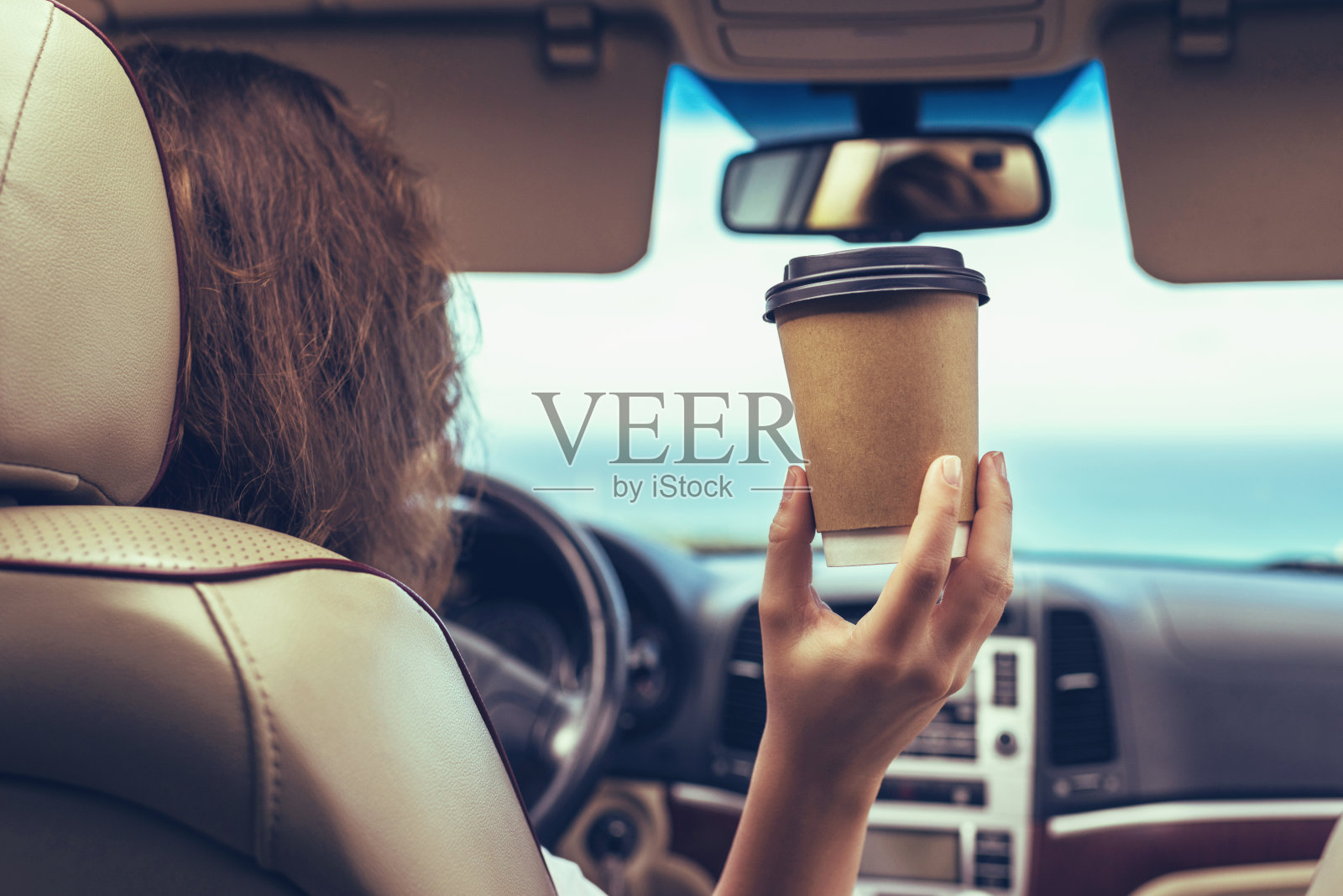 女司机开车时在车内喝咖啡纸杯。女孩放松在自动旅行沿着海洋热带海滩在背景。旅行的概念。后视图照片摄影图片