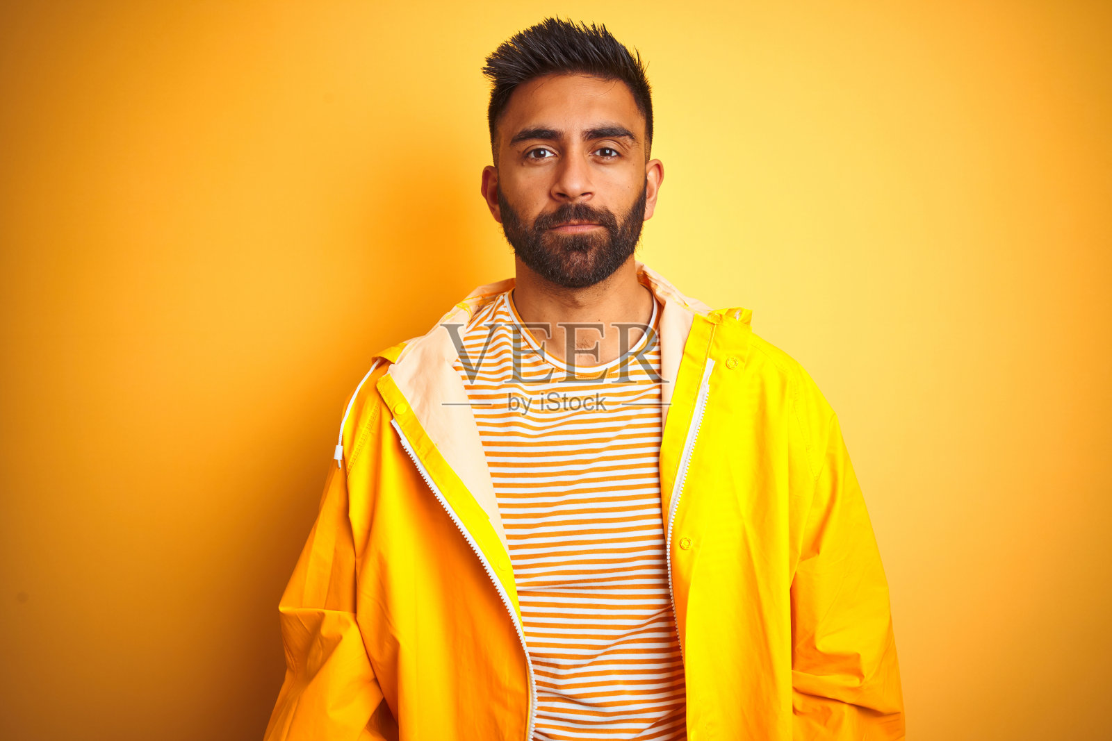 年轻的印度男子穿着雨衣站在孤立的黄色背景上，神情轻松，表情严肃。简单自然的看相机。照片摄影图片