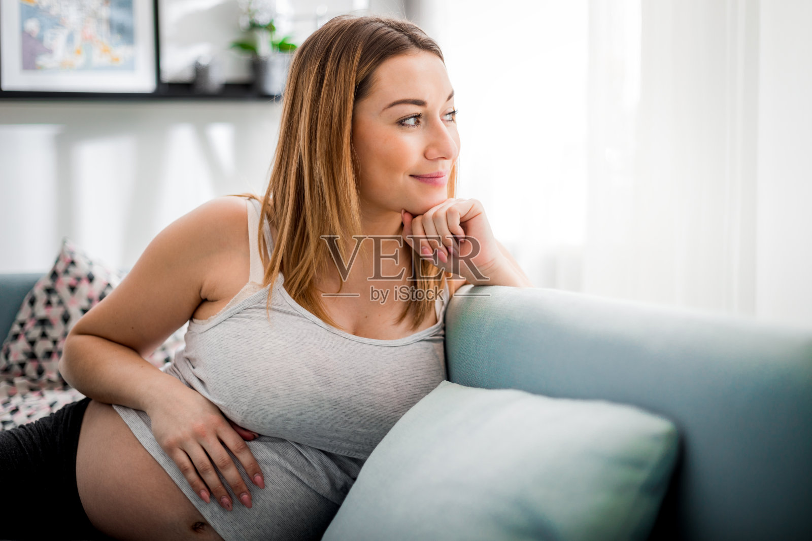 微笑的孕妇坐在沙发上，看着窗户，期待婴儿的概念照片摄影图片