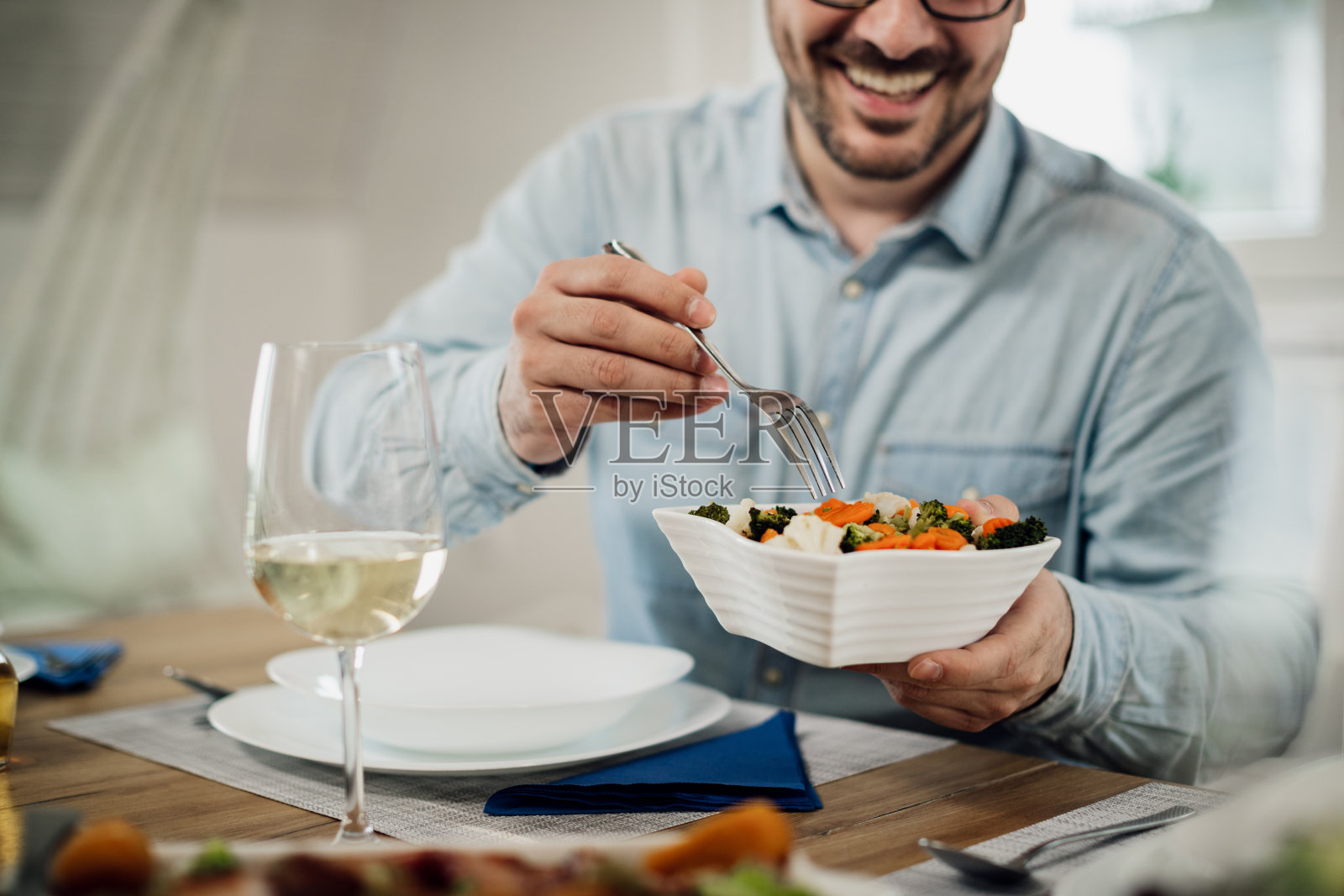 一个快乐的男人在餐桌上吃午餐时从碗里拿沙拉的特写。照片摄影图片