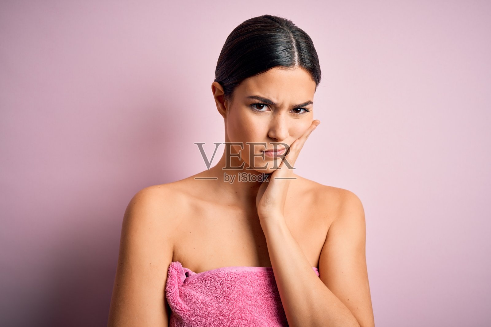 年轻美丽的女孩穿着浴巾淋浴后，站在孤立的粉红色背景思考看起来疲倦和抑郁的问题与交叉的胳膊。照片摄影图片