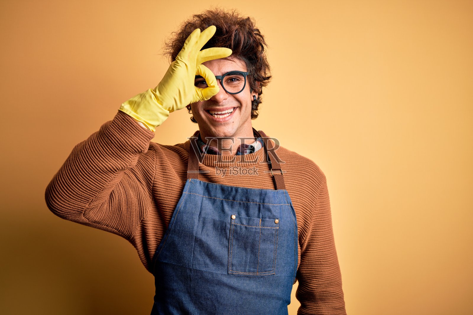 年轻英俊的清洁工穿着围裙和手套在孤立的黄色背景做ok手势与手微笑，眼睛透过手指与快乐的脸。照片摄影图片