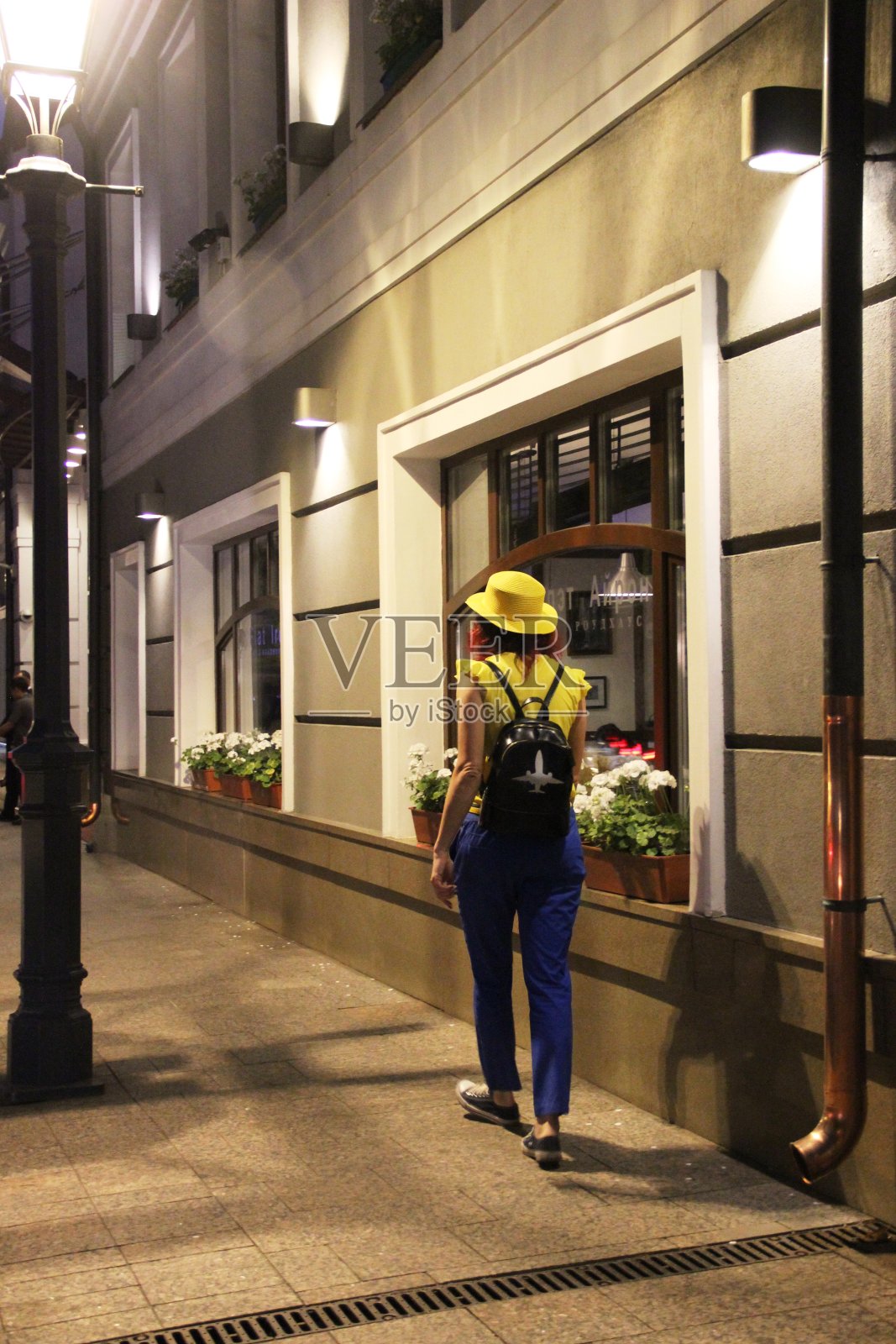 一个戴着黄色帽子的女人走开了，在远处沿着老城的夜街，背着黑白相间的背包。在莫斯科老城的女人在晚上走在城市街道上的背影照片摄影图片
