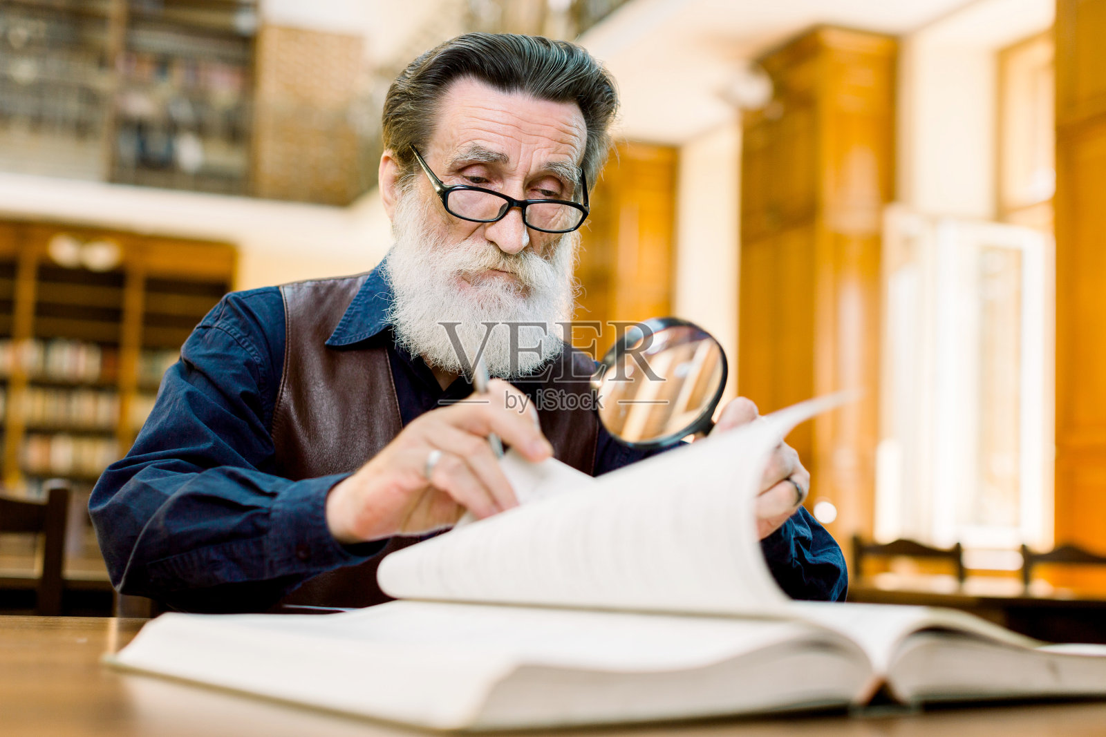 专注的上了年纪的时尚男老师、大学教授，戴着眼镜坐在旧图书馆里，一边翻书一边透过放大镜看照片摄影图片