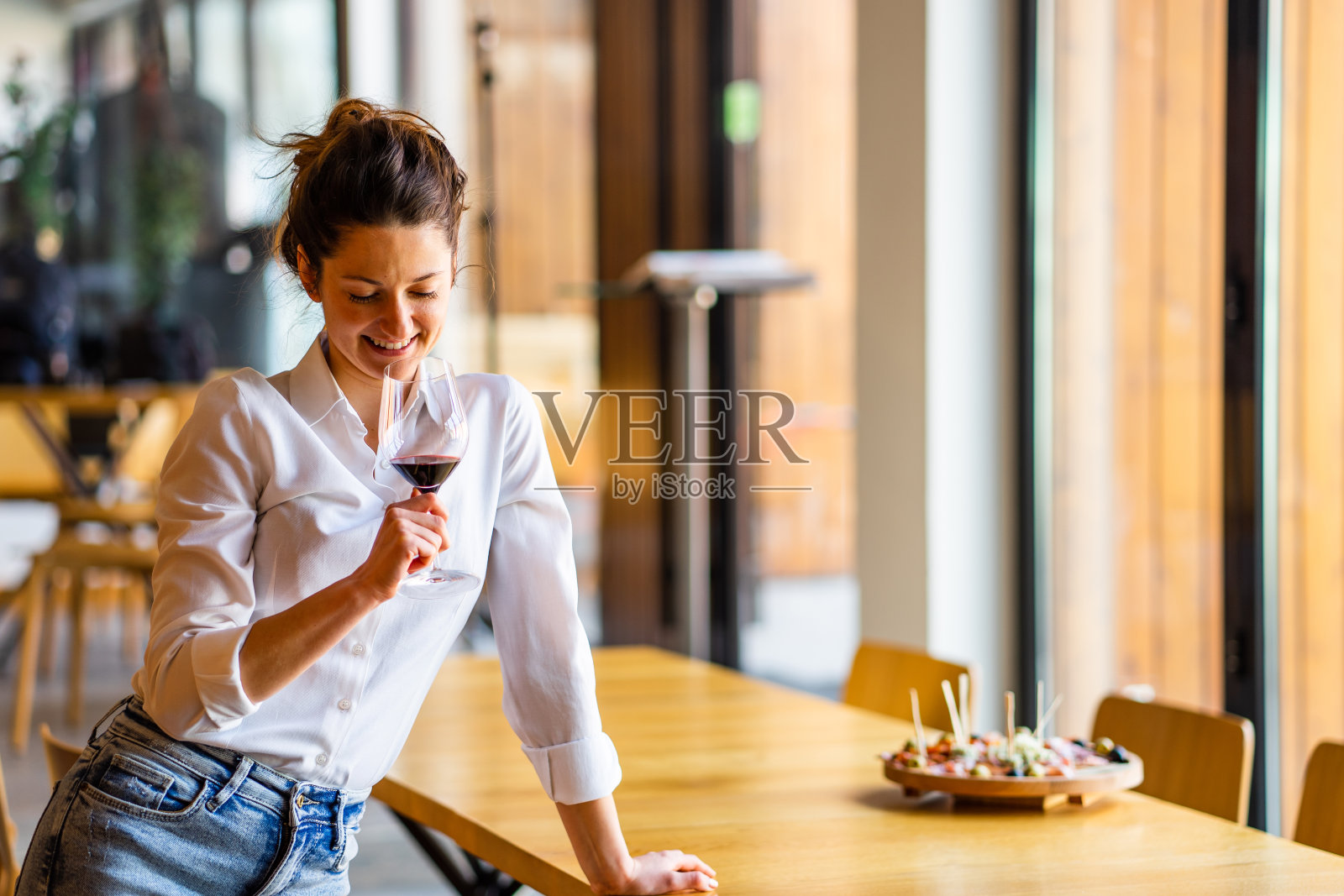 一个年轻美丽的白人妇女的肖像站在桌子靠握和看着在家里独自微笑着红葡萄酒穿着白衬衫正面视图照片摄影图片
