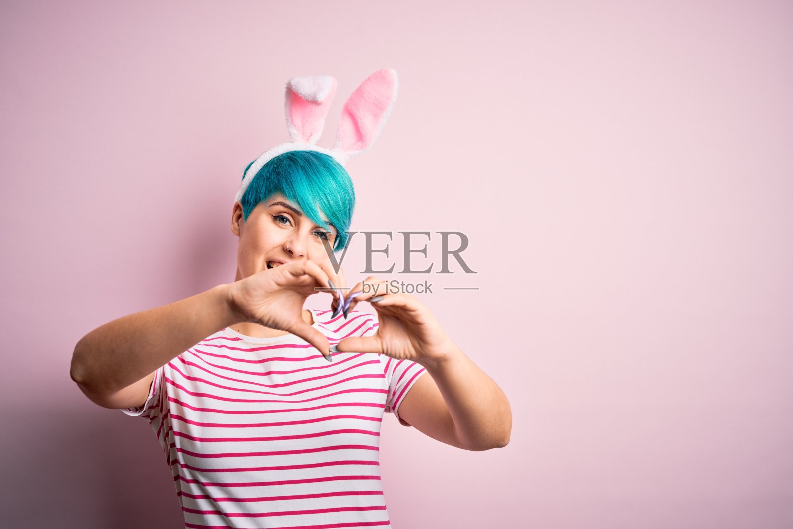 年轻女子与时尚的蓝色头发戴复活节兔子耳朵在粉红色的背景微笑做心形符号的手。浪漫的概念。照片摄影图片