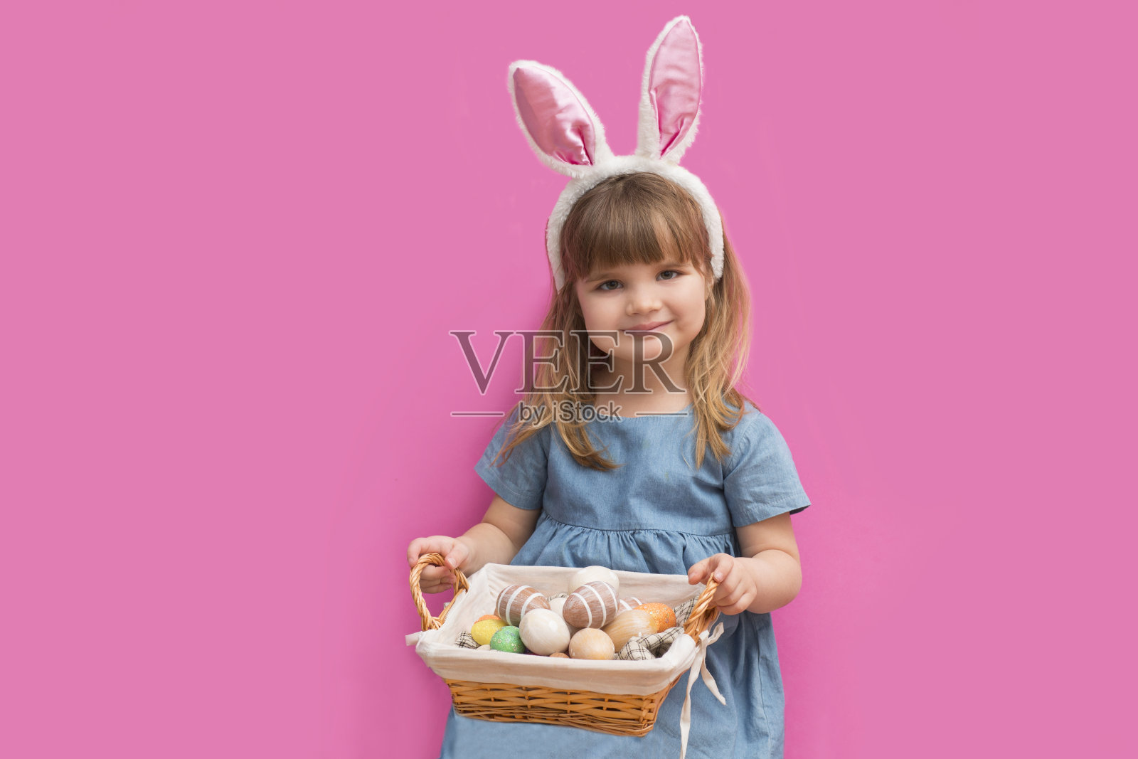 可爱的小女孩戴着兔子耳朵，拿着装有复活节彩蛋的篮子，在粉红色的背景上摆姿势。复活节快乐!照片摄影图片