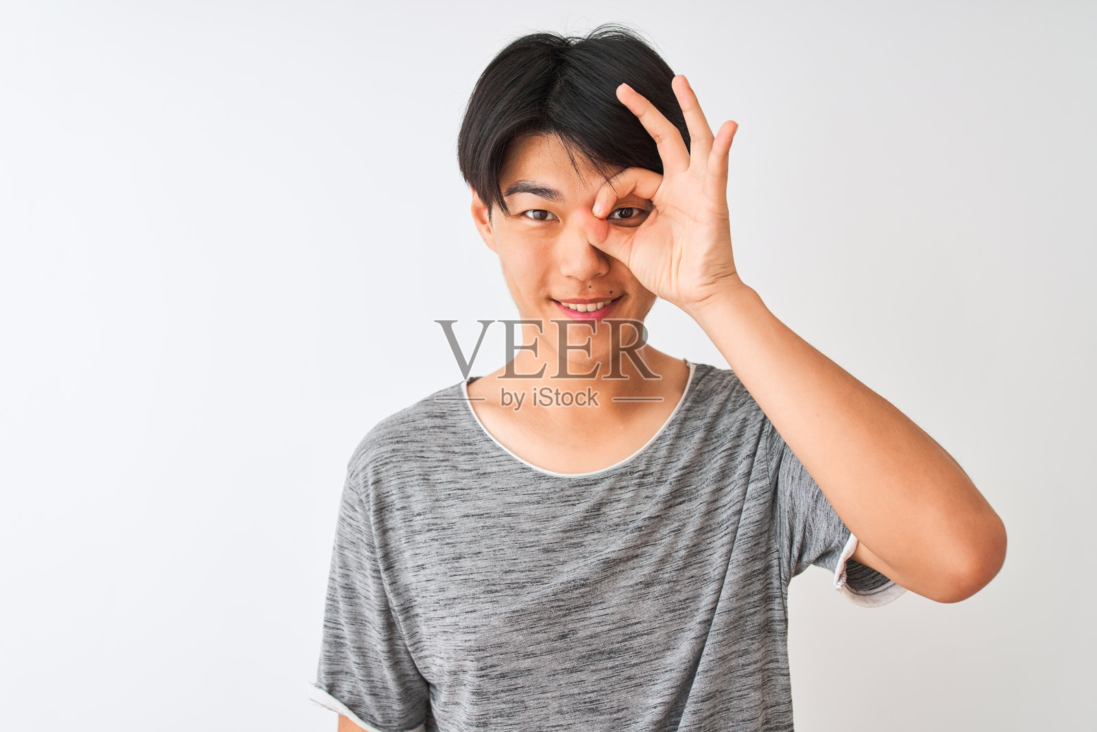 年轻的中国男子穿着休闲t恤站在孤立的白色背景与快乐的脸微笑做ok手势与手在眼睛透过手指看照片摄影图片