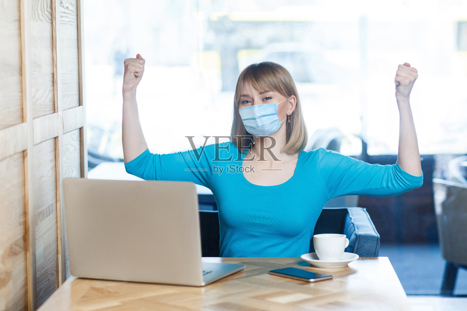 我是坚强的。一个穿着蓝色t恤戴着医用外科口罩的年轻女子的肖像正坐在那里看着摄像机，展示着她的二头肌，很满意。照片摄影图片