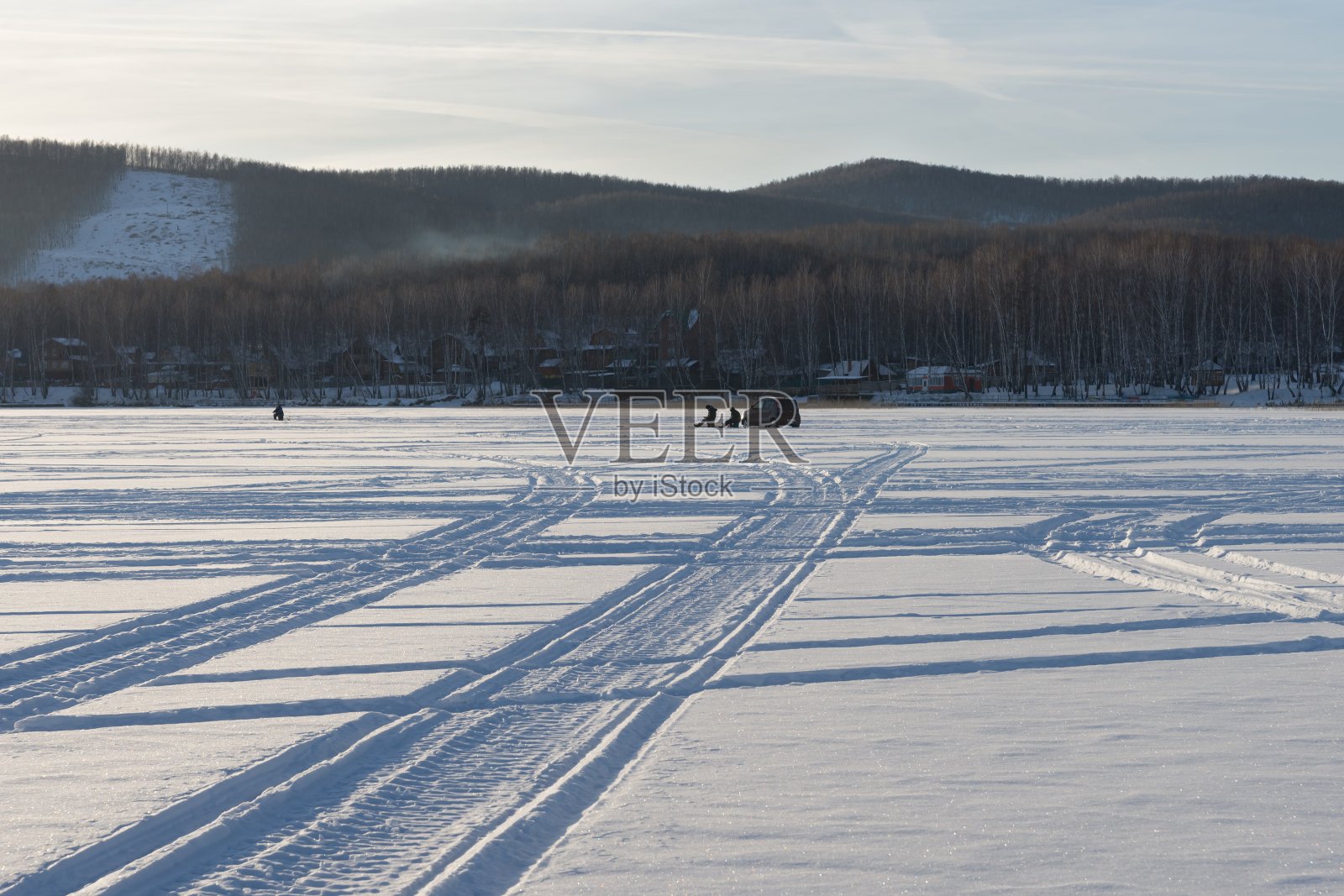 车轮的痕迹从雪原的湖到渔民背对海岸的背景。照片摄影图片