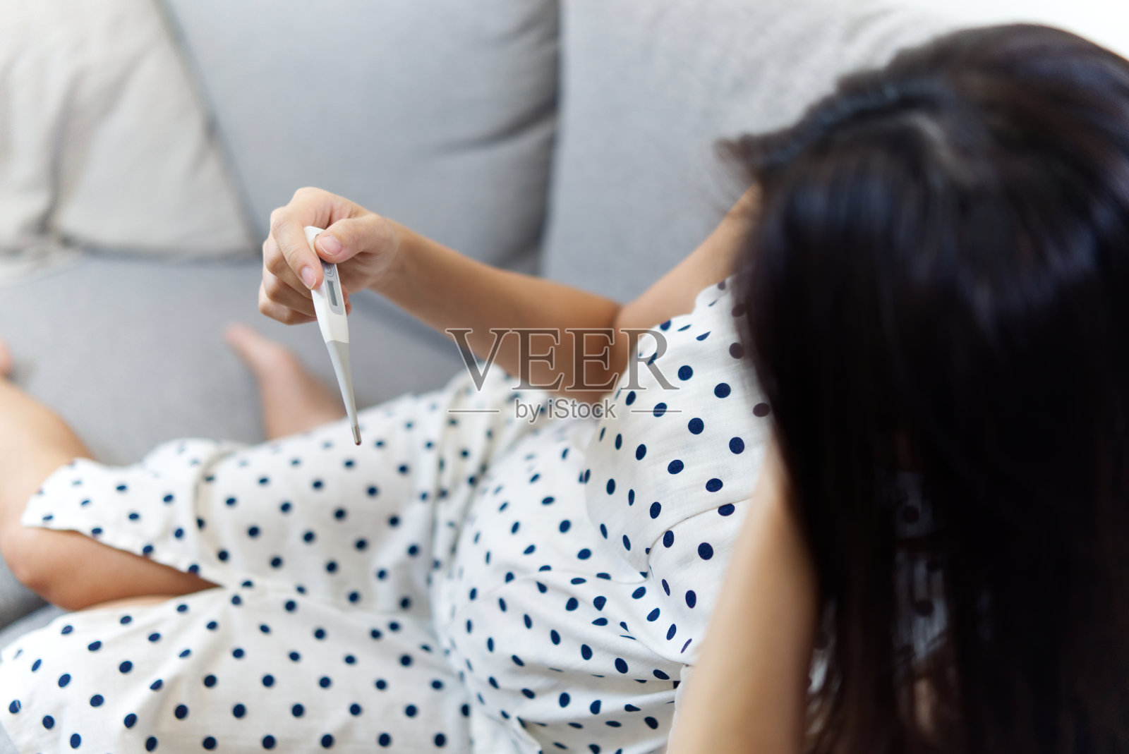 疾病或疾病的怀孕妇女的概念。亚洲怀孕女孩发烧了，她坐在家里的沙发上看温度计上的温度，感觉很不好。照片摄影图片