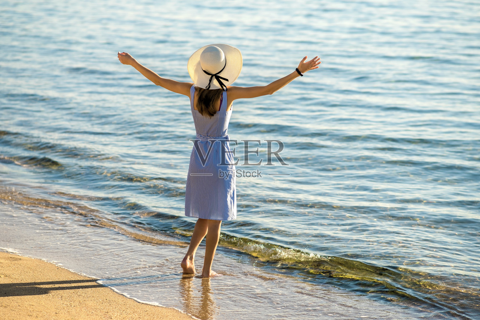 年轻女子戴着草帽，穿着裙子，站在海边空旷的沙滩上举手。孤独的女游客在度假中看着平静的海面上的地平线。照片摄影图片