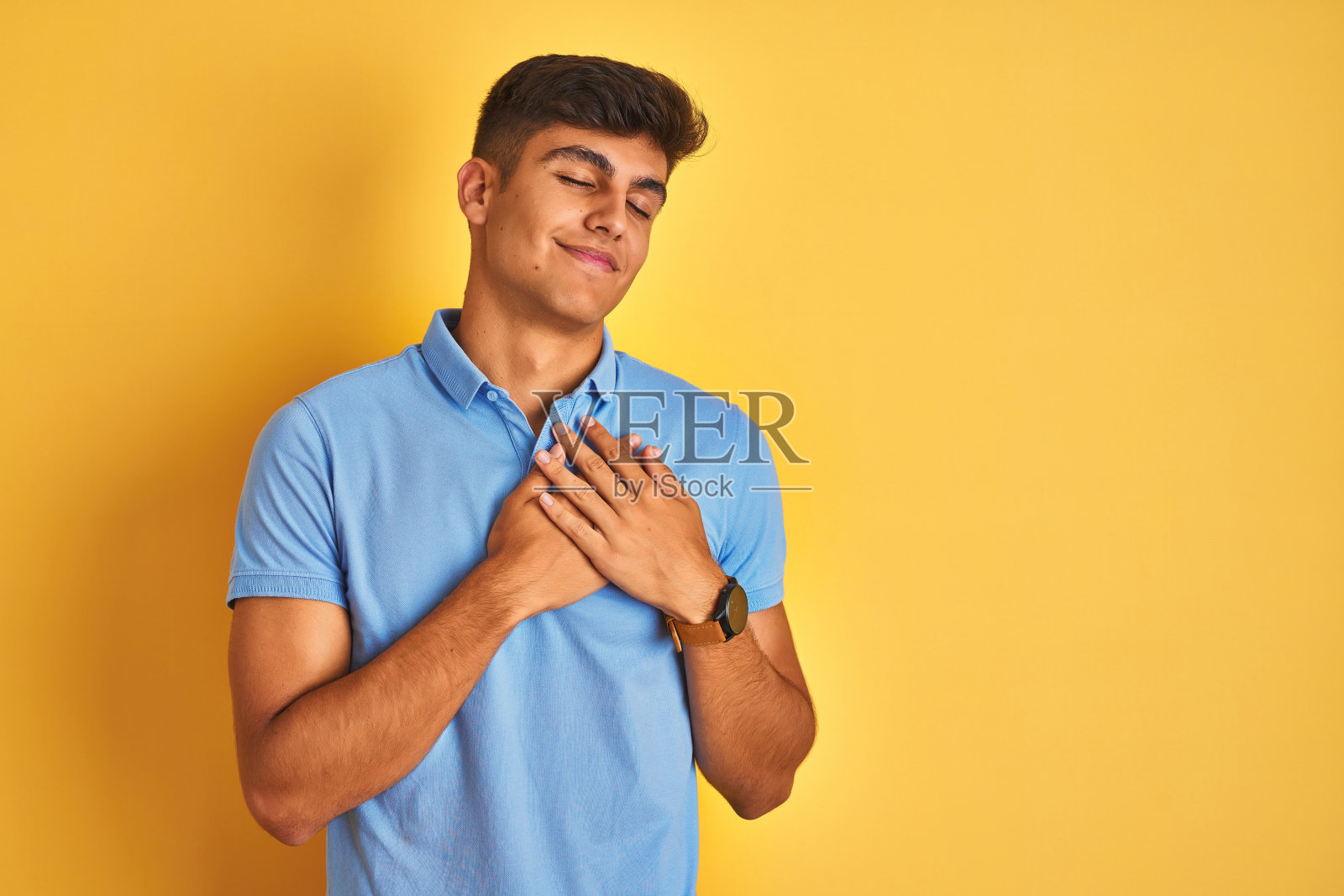 年轻的印度男子穿着蓝色马球站在孤立的黄色背景微笑，双手放在胸前，闭上眼睛和感激的手势在脸上。健康的概念。照片摄影图片
