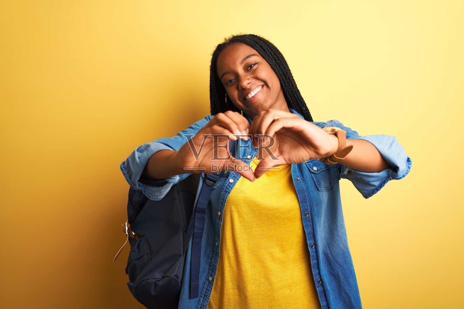 非洲裔美国女学生背着背包站在孤立的黄色背景微笑在爱显示心形符号和形状的手。浪漫的概念。照片摄影图片