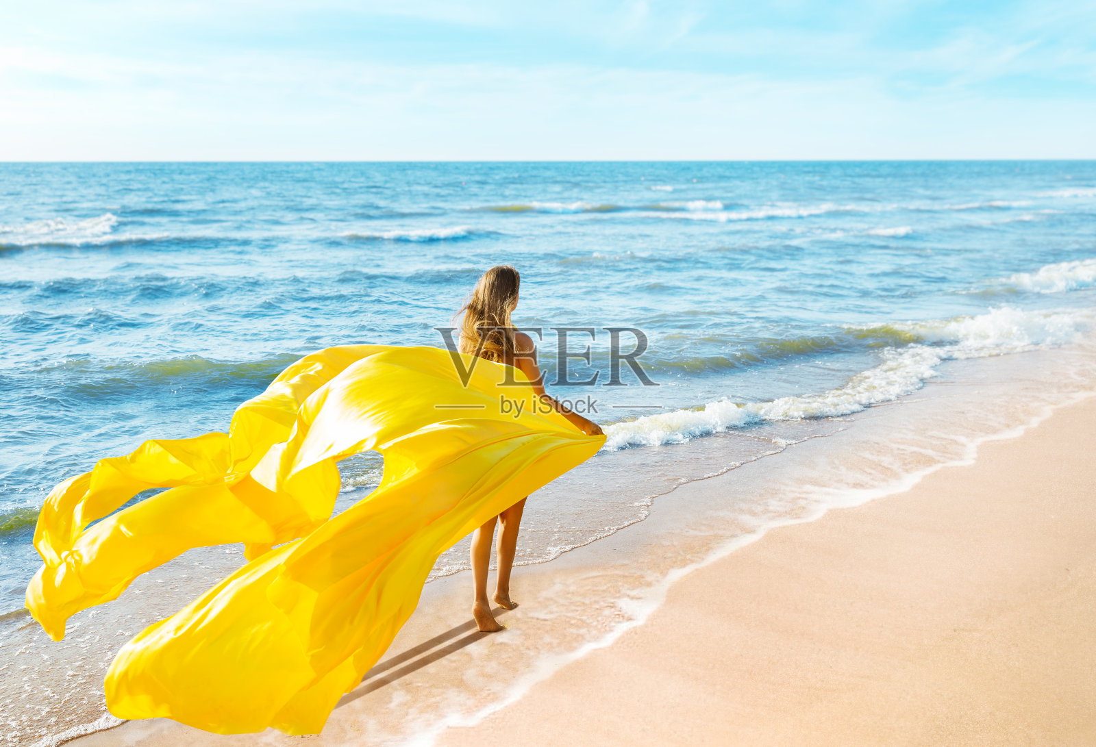 时尚模特穿着黄色飘动的连衣裙走在沙滩上，女人后视镜，丝绸在风中飘扬照片摄影图片