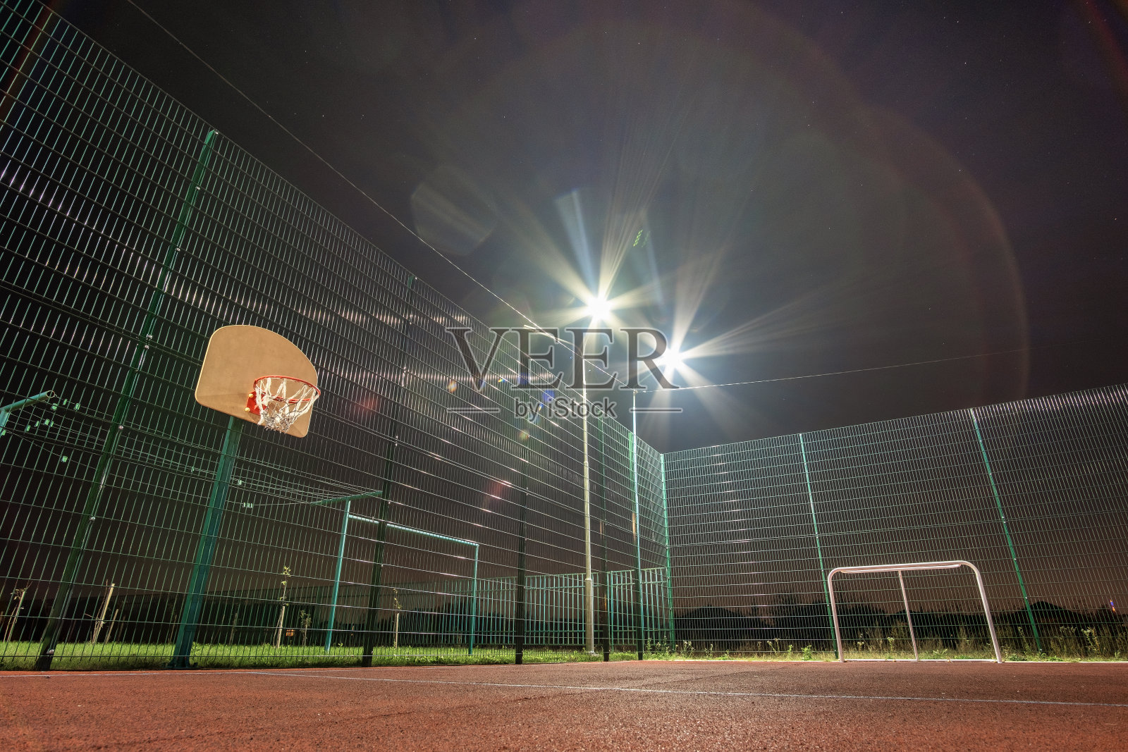 户外迷你足球场和篮球场，球门和篮筐环绕着高防护网，在夜晚被聚光灯照亮。照片摄影图片
