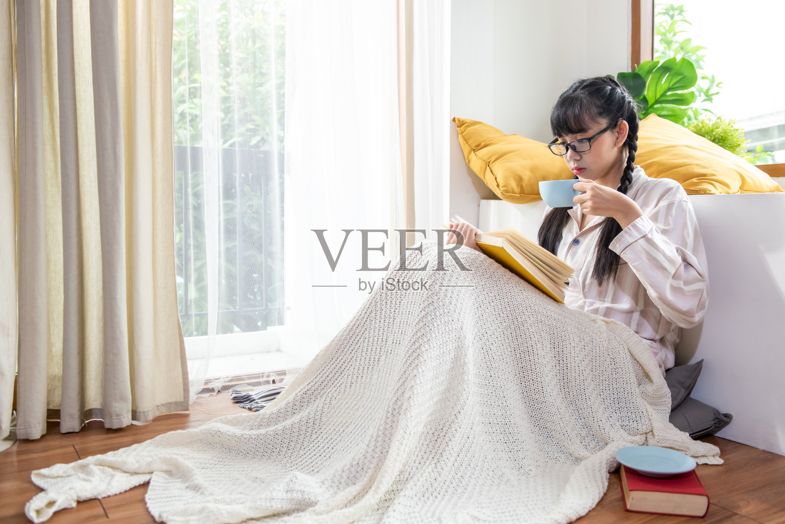 年轻美丽的亚洲少女穿着睡衣，戴着眼镜，独自坐在卧室地板上，一边喝着热咖啡或茶一边看书。用在家上学的方式让女学生放松。照片摄影图片