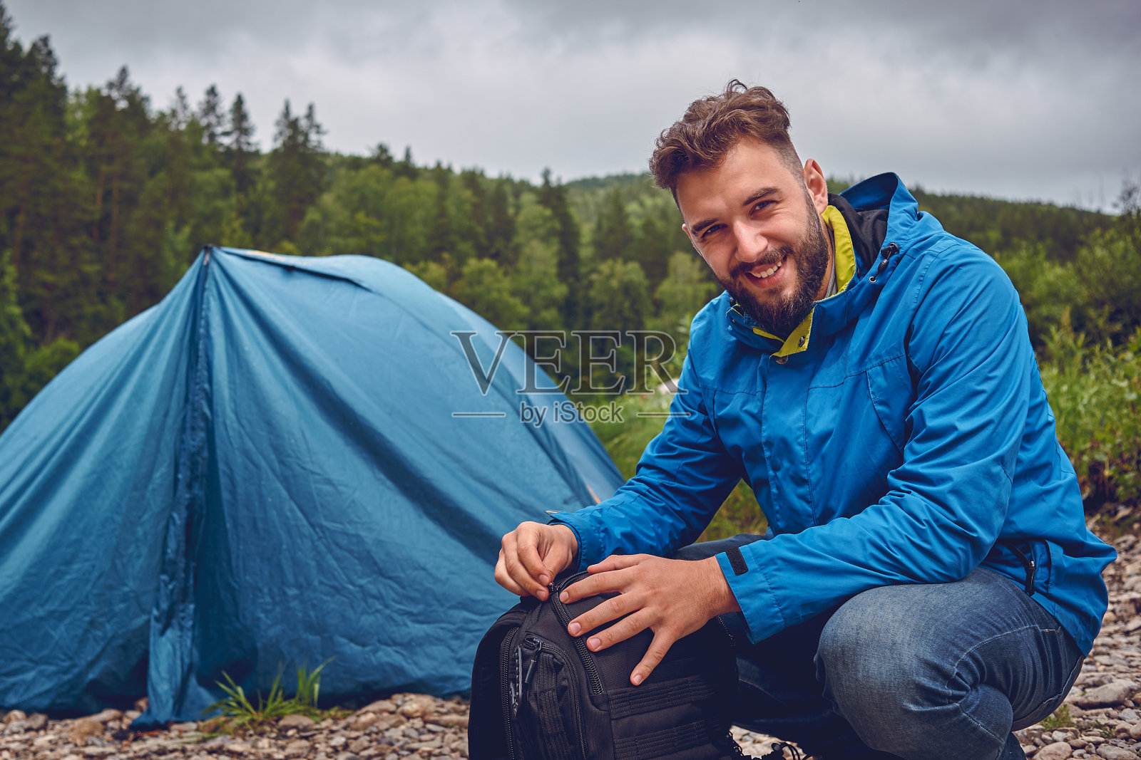 一个留着胡子的年轻人背着背包，在户外露营，背景是一个旅游帐篷照片摄影图片