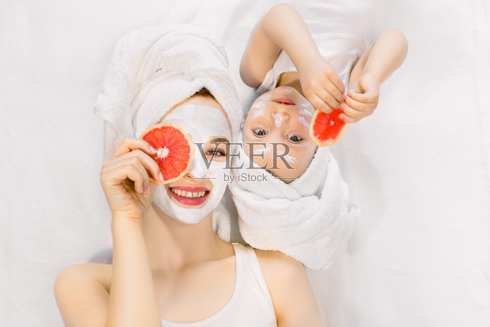 年轻漂亮的妈妈和她两岁的小女儿头上裹着毛巾，一起躺在床上，做着水疗护肤程序，敷着面膜，拿着眼睛附近的葡萄柚照片摄影图片