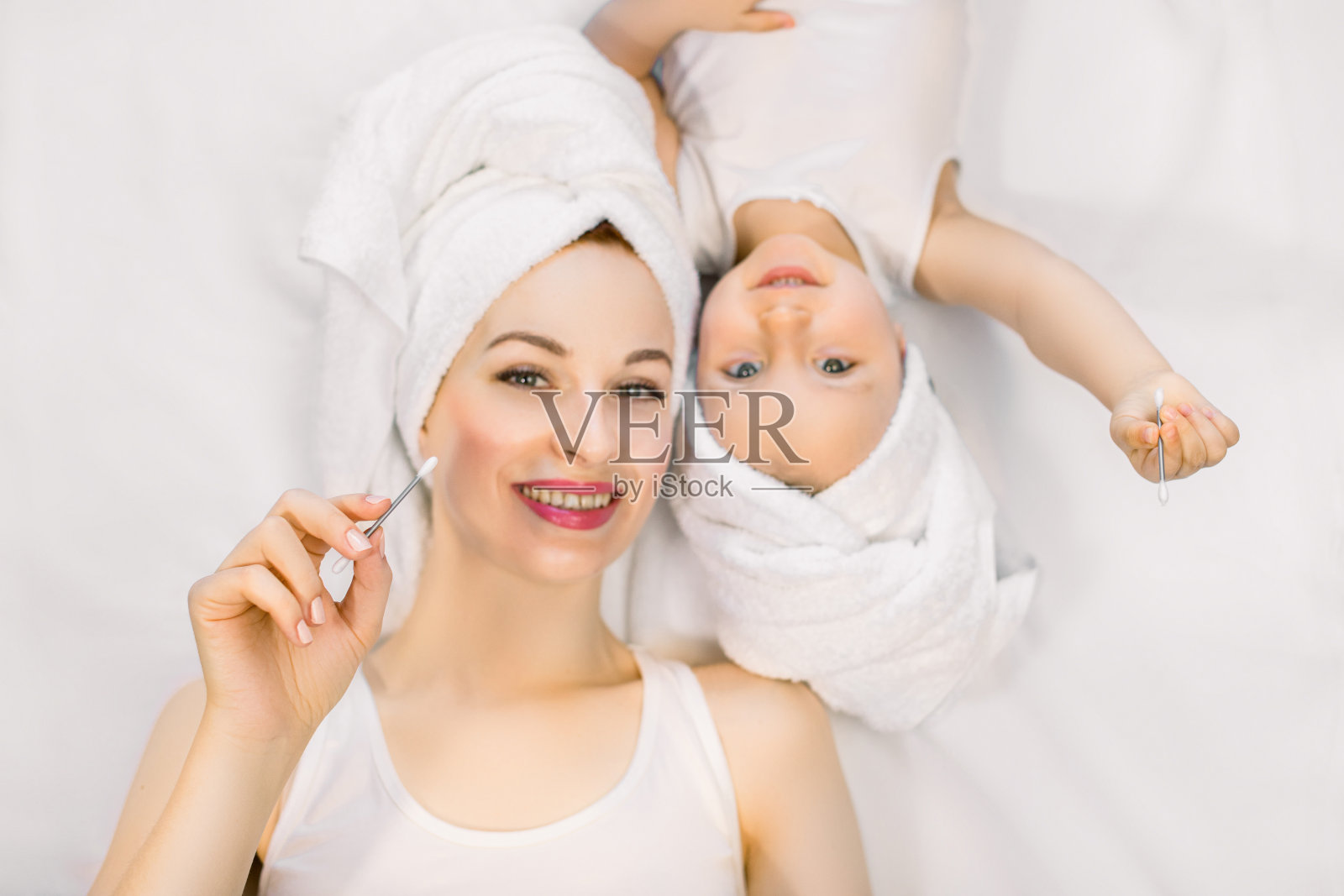 年轻积极迷人的妈妈和可爱的小宝贝女儿头顶毛巾，一起躺在床上，手里拿着耳棒。家庭沐浴美容。选择性地关注手照片摄影图片
