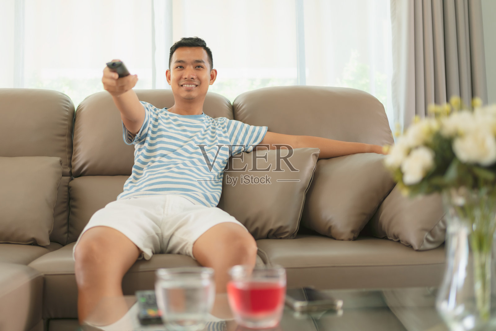 亚洲年轻人坐在沙发上，拿着遥控器，在家里的客厅看电视。照片摄影图片