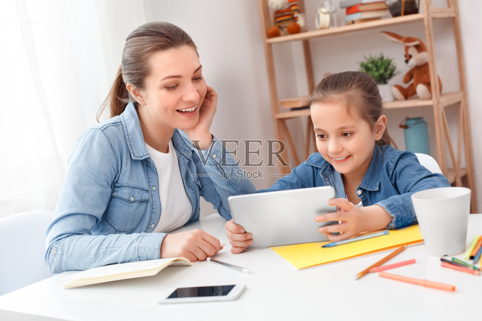 家庭的概念。母亲和女儿一起玩电子平板电脑，一起呆在家里，做作业，阅读新的儿童书籍。小女孩笑着跟导师姐姐玩。两个人坐在桌边。可爱的照片摄影图片
