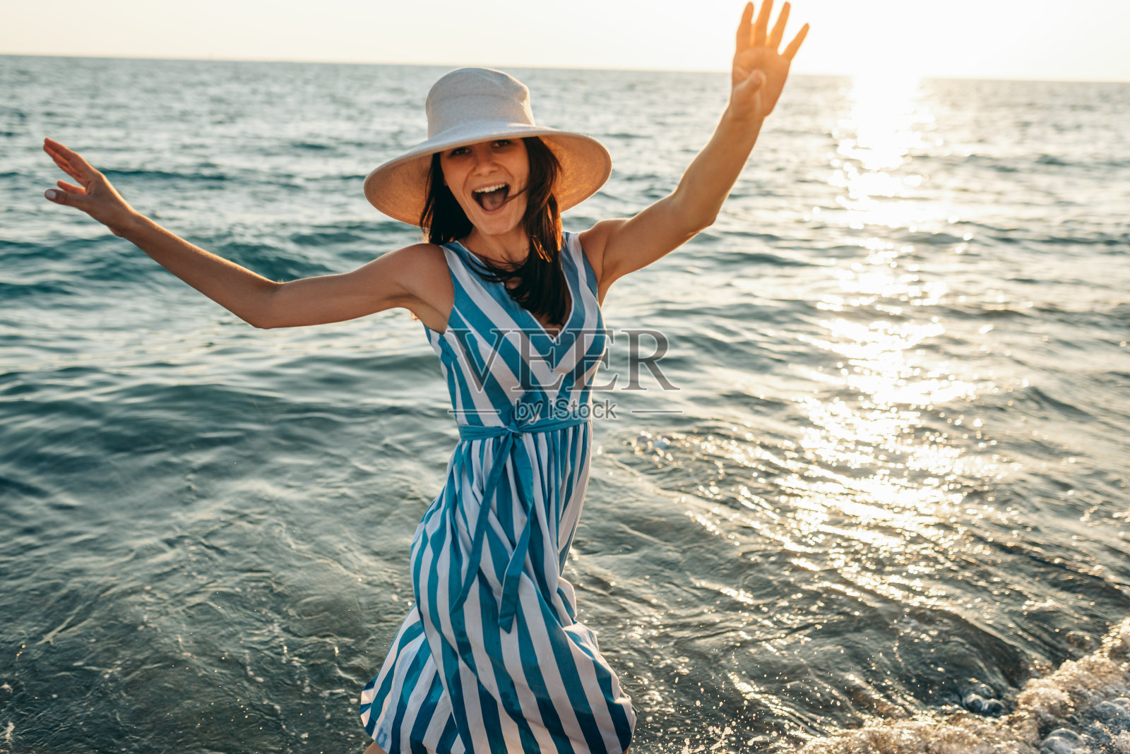 一个快乐的年轻女人的肖像微笑着双臂向上，戴着白色的帽子在头上，和条纹的衣服，走在海边的海滩上日落的阳光。旅行的概念。照片摄影图片