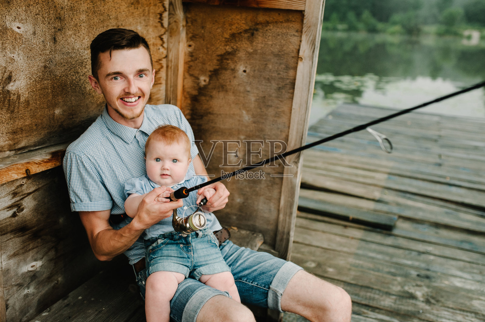 父亲和女儿坐在木码头上，在湖的背景钓鱼。一家人在池塘钓鱼。家庭度假钓鱼。夏天，年轻的爸爸带着孩子在河边户外。照片摄影图片