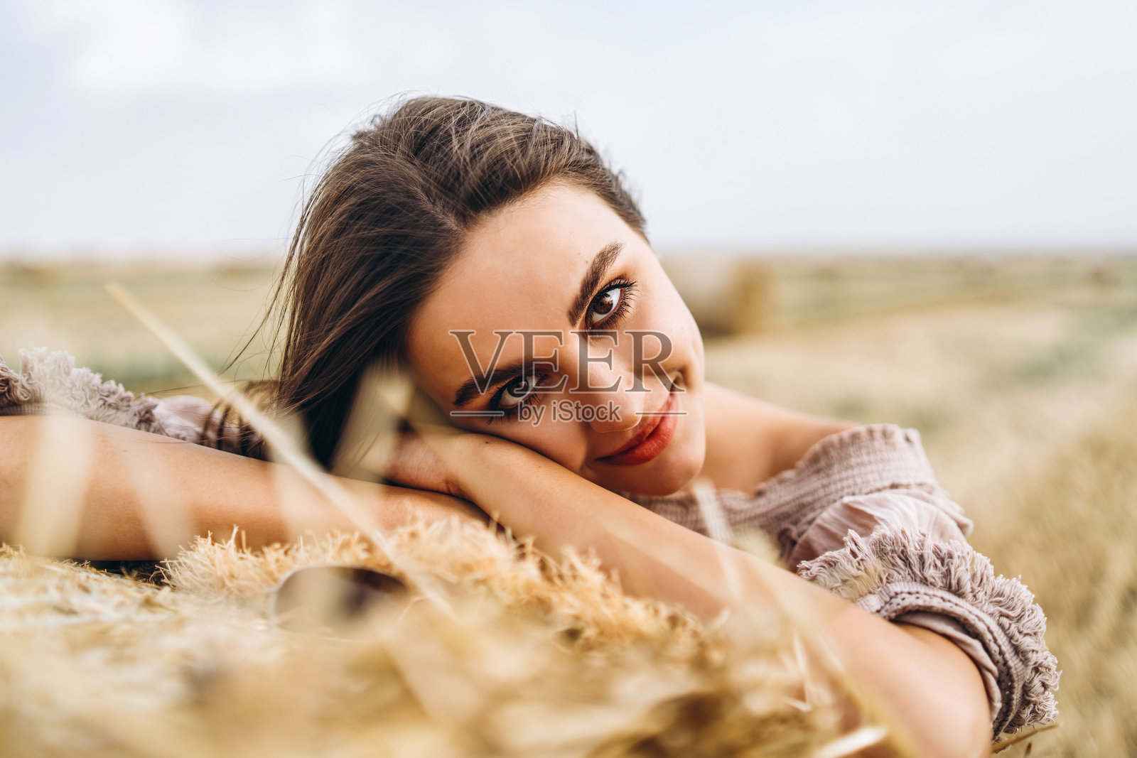 闭着眼睛微笑的美丽女人的特写。黑发女人靠在一捆干草上。背景是一片麦田照片摄影图片