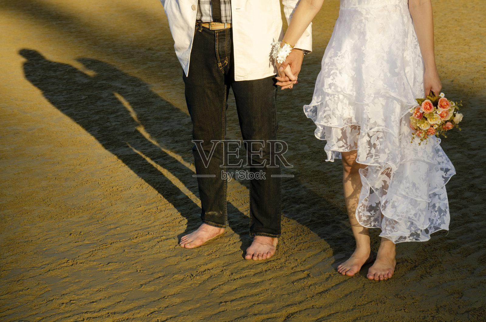 夕阳下的沙滩上，新娘和新郎手牵着手，穿着美丽的婚纱，长长的影子，新婚夫妇正享受着彼此，手持新娘花束。照片摄影图片