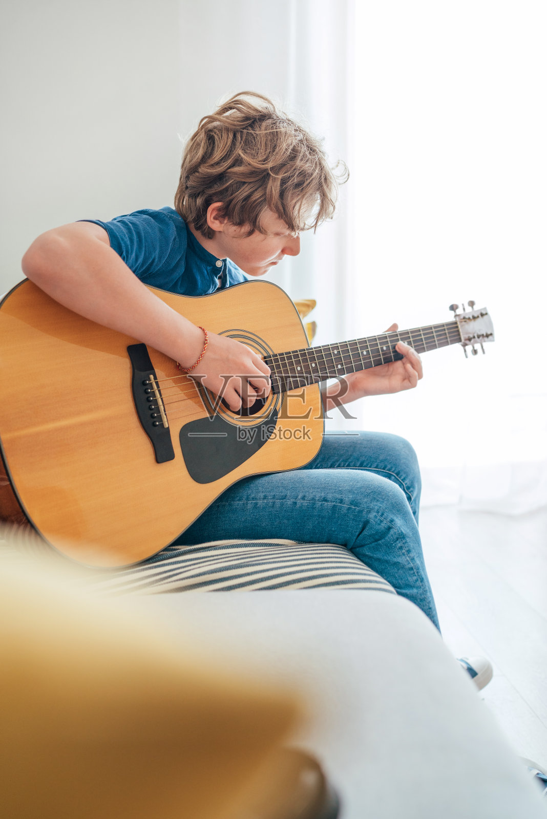 一个十几岁的男孩，穿着休闲牛仔裤和衬衫，弹着原声吉他，坐在家里客厅舒适的沙发上。音乐教育理念形象。照片摄影图片