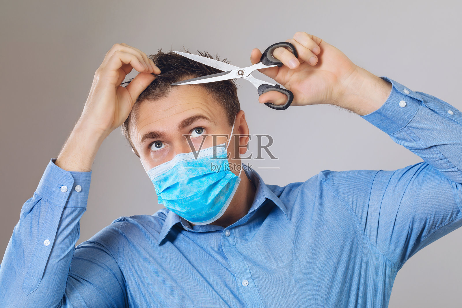 一名戴着医用口罩的男子试图在居家隔离的同时，为自己独立提供美容服务。冠状病毒导致的封闭美容院的概念照片摄影图片