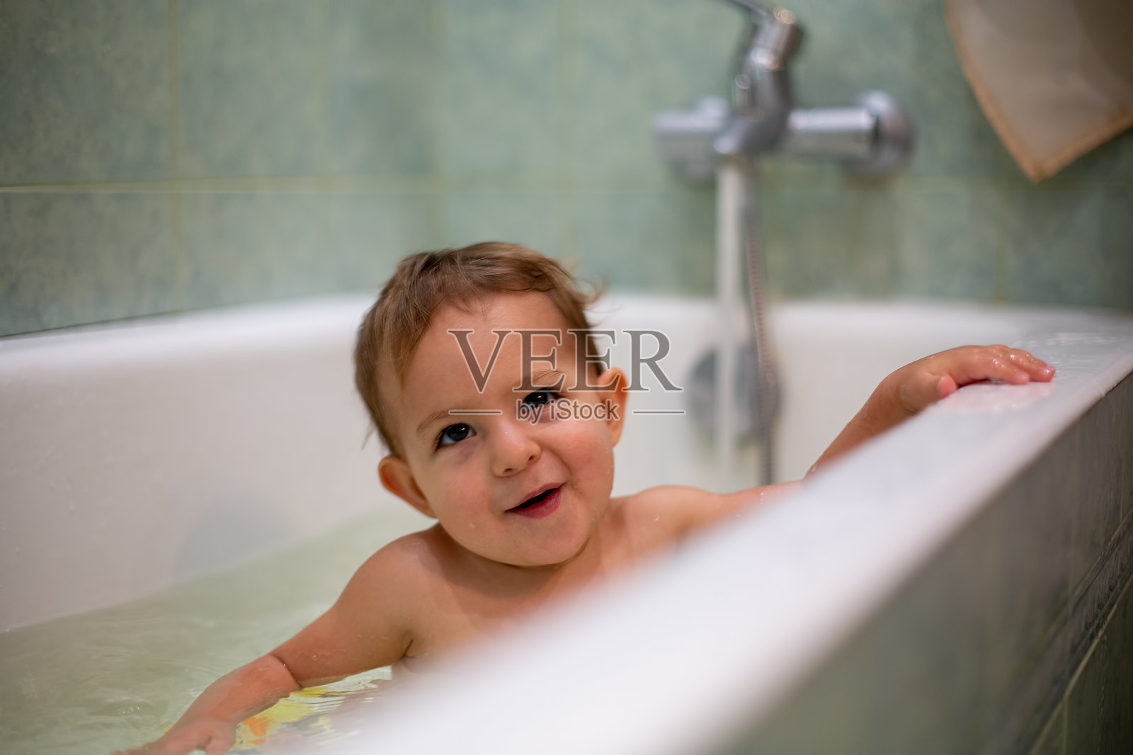 可爱的白种婴儿正在洗澡，调皮地微笑，抬头看，休息在浴缸的一边。水飞溅，背景是一间模糊的绿色浴室。特写,软焦点照片摄影图片