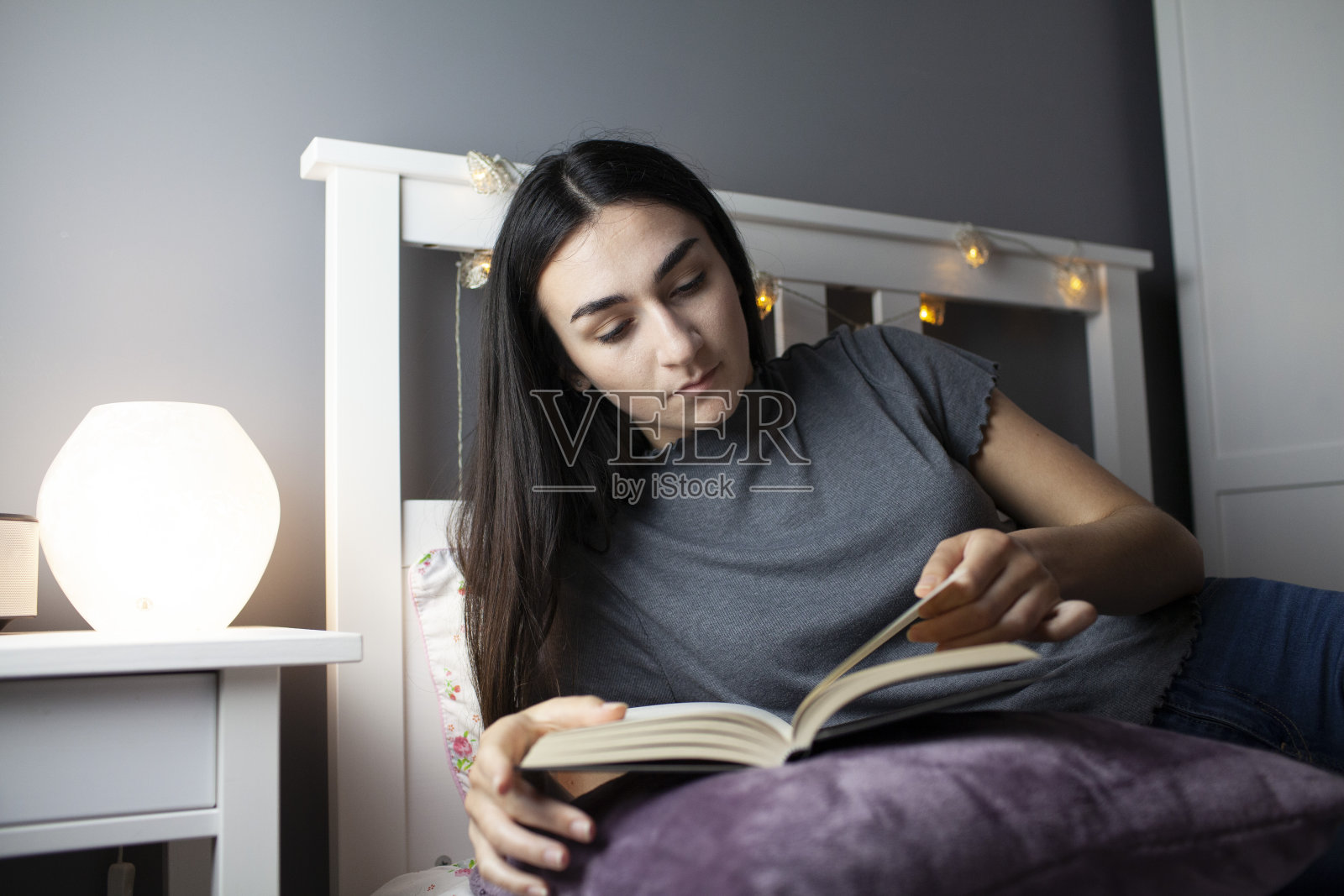 一个十几岁的女孩在她卧室的床上看书，床边的灯照片摄影图片