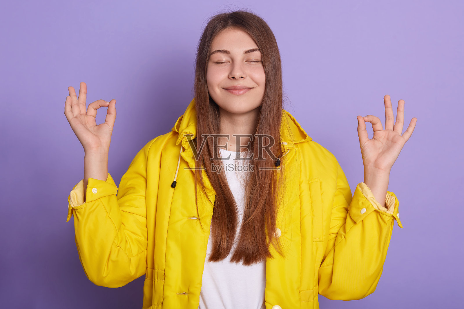 特写的快乐微笑的女人摆姿势闭上眼睛，保持食指和拇指在一起，穿着黄色夹克，放松的女孩有深色头发，站在淡紫色工作室的墙。照片摄影图片
