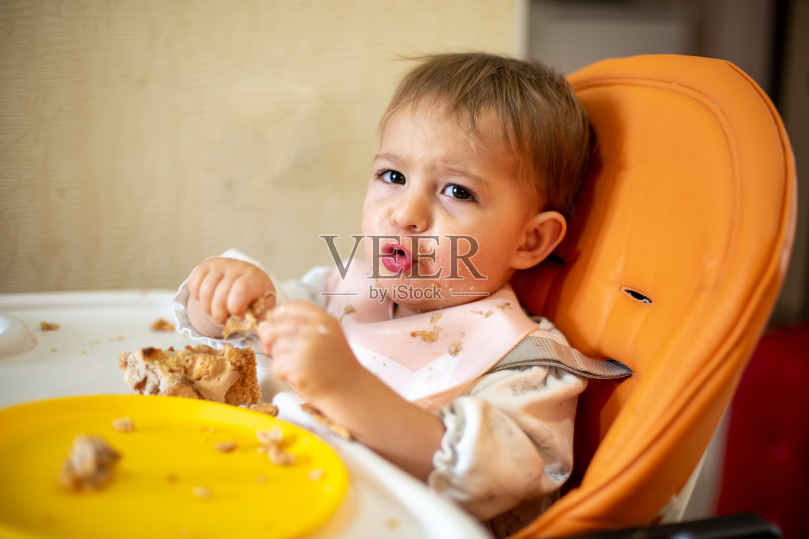 一个可爱的婴儿坐在一个橙色的儿童座椅和一张桌子上，他的手和脸都很脏，他看着镜头，做着鬼脸。桌子上有面包屑和一个橙色的盘子。特写，前视图，软焦点，模糊背景照片摄影图片