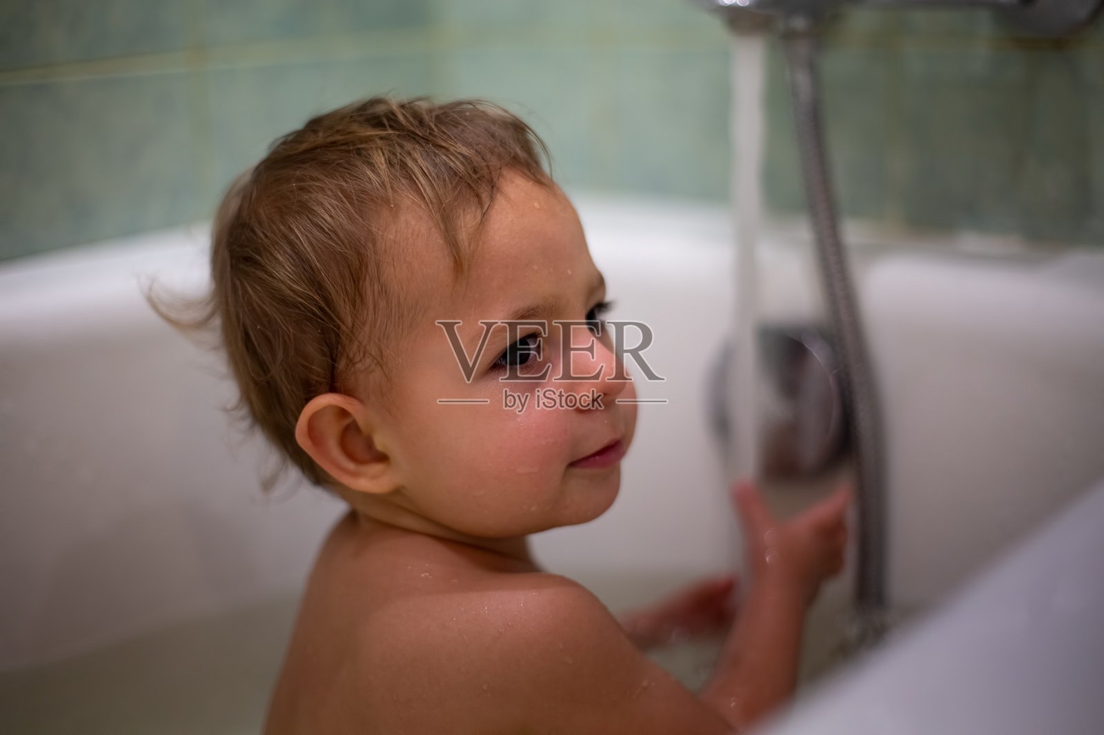一个可爱的白种婴儿在洗澡时一边玩水一边把头扭向一边。特写，柔焦，背景中有一个水龙头，水是模糊的照片摄影图片