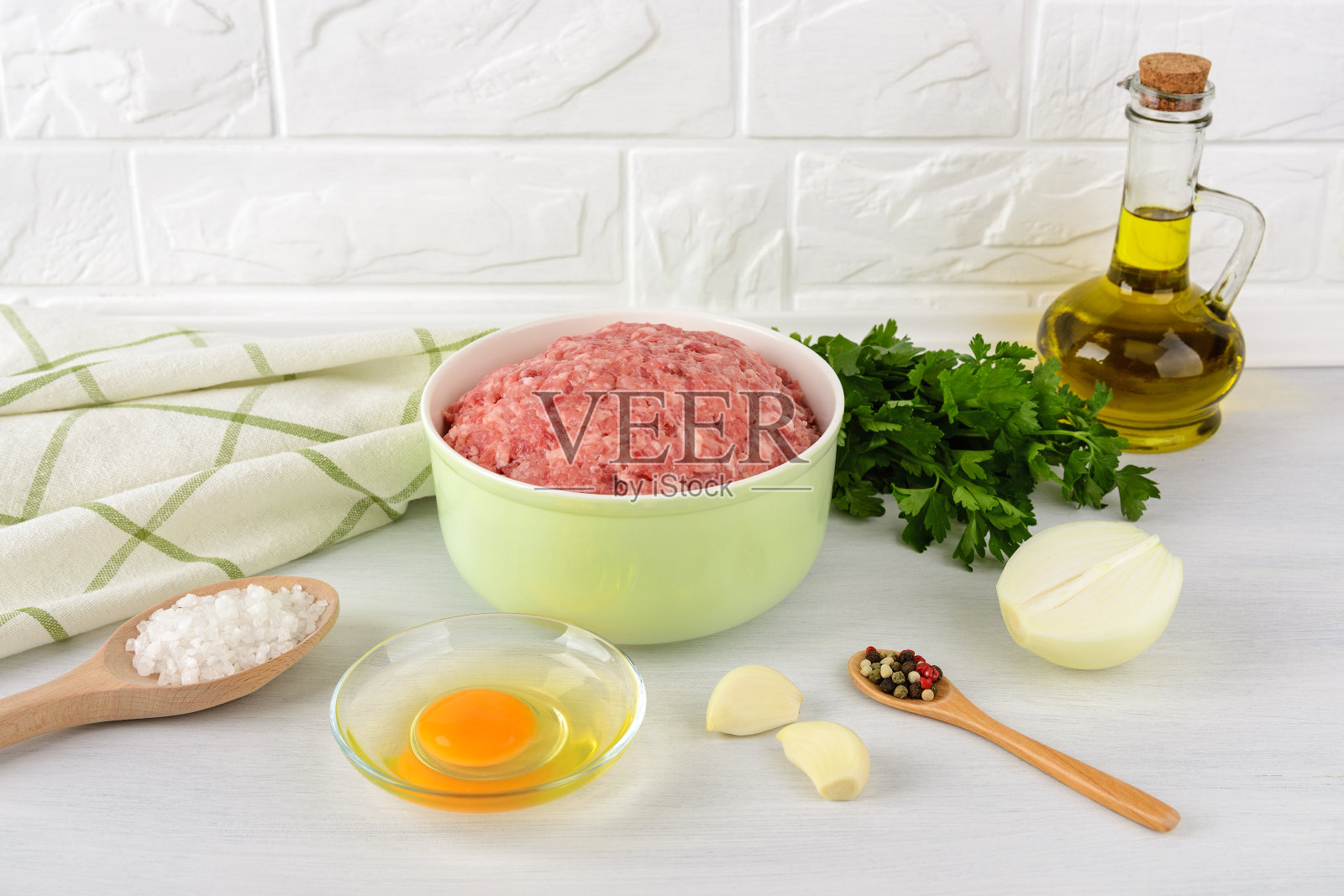 新鲜的肉末放在一个绿色的碗里，鸡蛋，洋葱，大蒜，盐和胡椒放在厨房的桌子上。肉片或肉丸的原料在家做饭。照片摄影图片