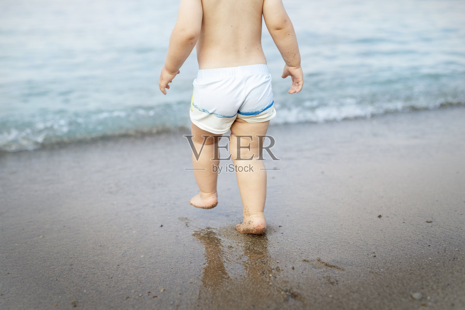 后视图可爱可爱的单独的小高加索蹒跚学步的男孩在游泳短裤走过蓝色清澈的水海浪热带海洋或海洋海滩。假期和旅行与小孩子的概念照片摄影图片