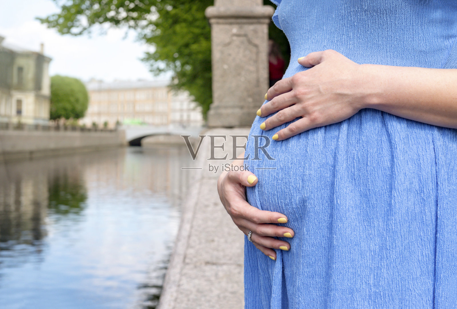 一名身穿蓝色连衣裙的白人孕妇腹部特写在江畔的城市，双手拥抱着她的腹部，黄色的指甲照片摄影图片