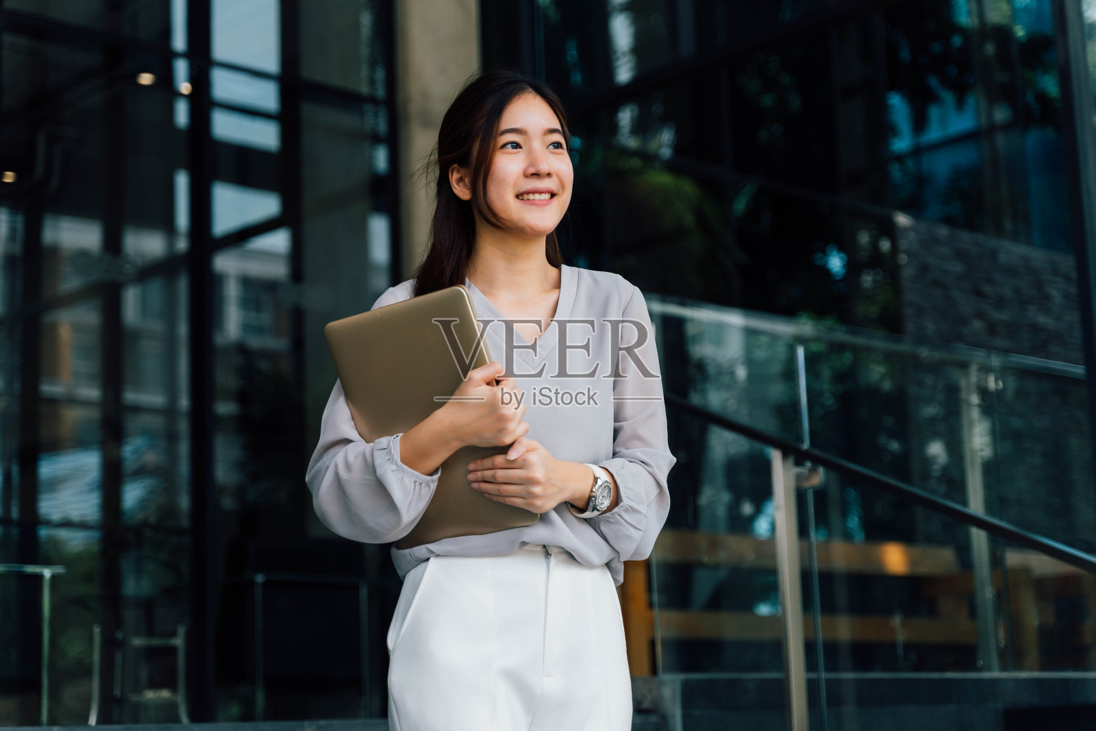 一位年轻成功的商业女性微笑着拿着一台笔记本电脑。职业女性工作者站在公司现代办公室和窗户在休闲灰色的样子。-有拷贝空间照片摄影图片