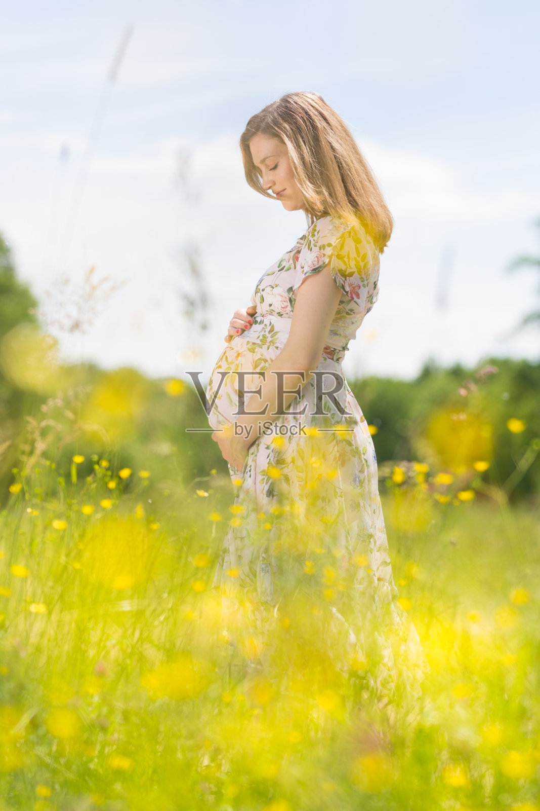 美丽的孕妇穿着白色的夏装，在满是黄色花朵的草地上。照片摄影图片