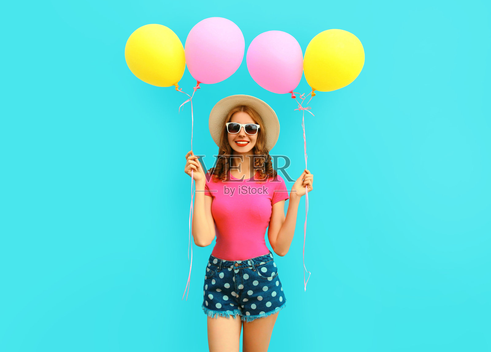 夏天彩色的愉快的图像快乐的年轻女子与黄色的粉红色气球有乐趣穿着短裤和草帽在蓝色的墙壁背景照片摄影图片