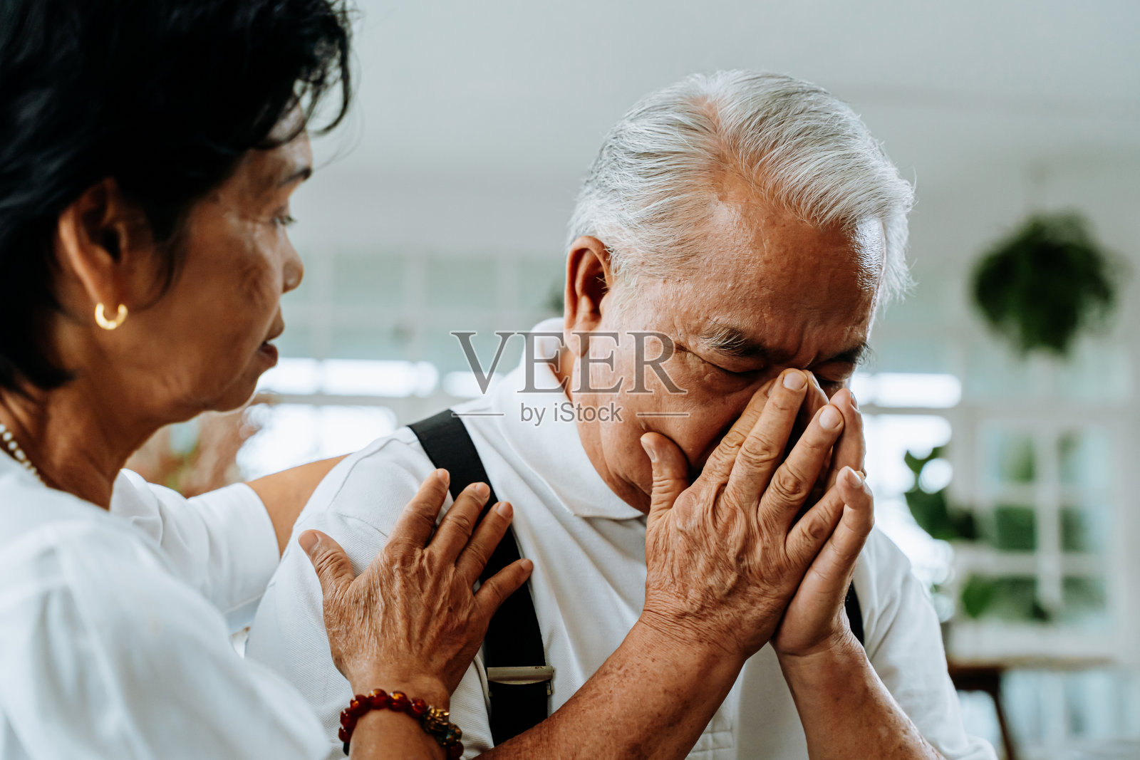 不快乐的退休亚洲老夫妇坐在沙发上，而老妻子安慰她的悲伤的丈夫在家里。照片摄影图片
