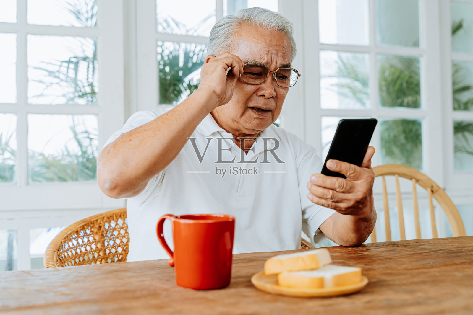 年长的亚洲男性有视力问题，在家里吃早餐时戴着眼镜盯着智能手机看信息。在室内视力模糊的老人照片摄影图片