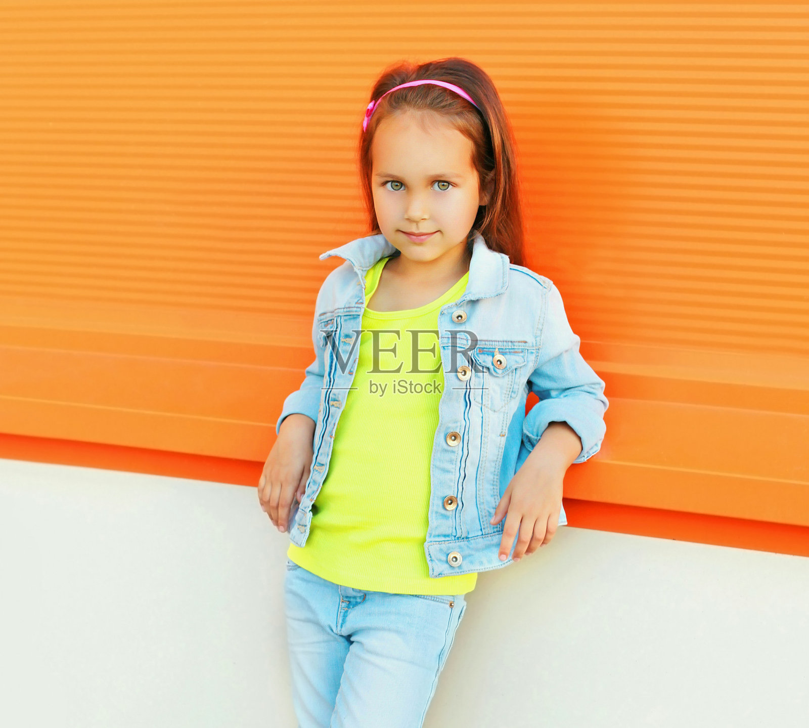 一个漂亮的小女孩的肖像，戴着太阳镜，牛仔裤夹克在城市街道上橙色的背景照片摄影图片